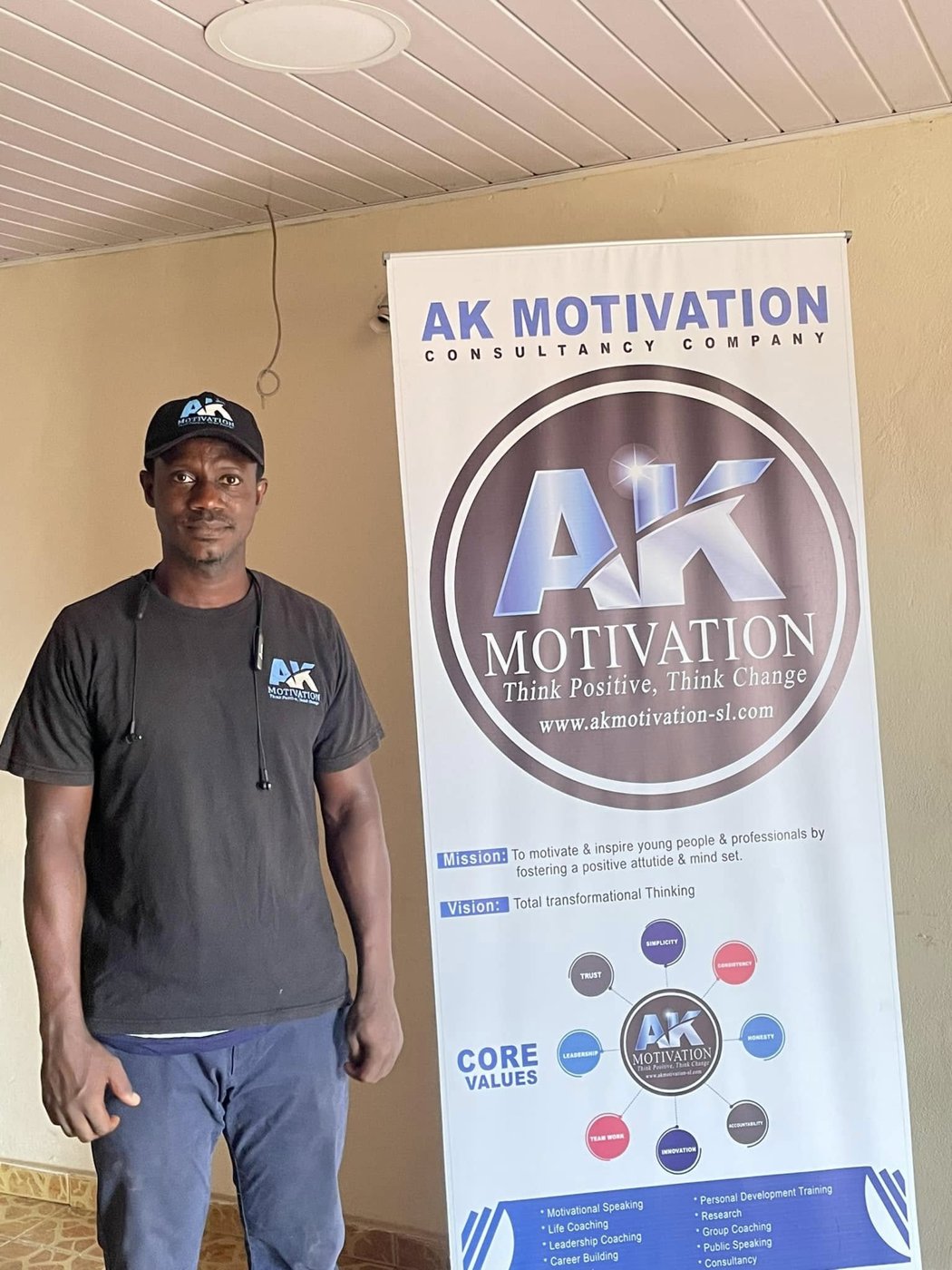 Abdulai standing beside the AK Motivation Consultancy Company banner showing its mission, vision and core values
